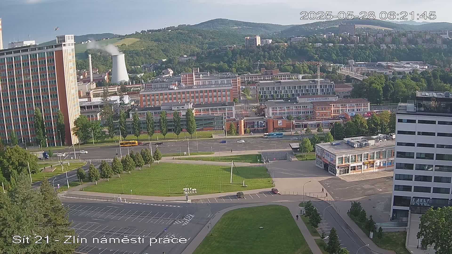 A high-angle, long shot on a partly sunny day shows a city square with several buildings, including a large high-rise, surrounded by green space and roads.