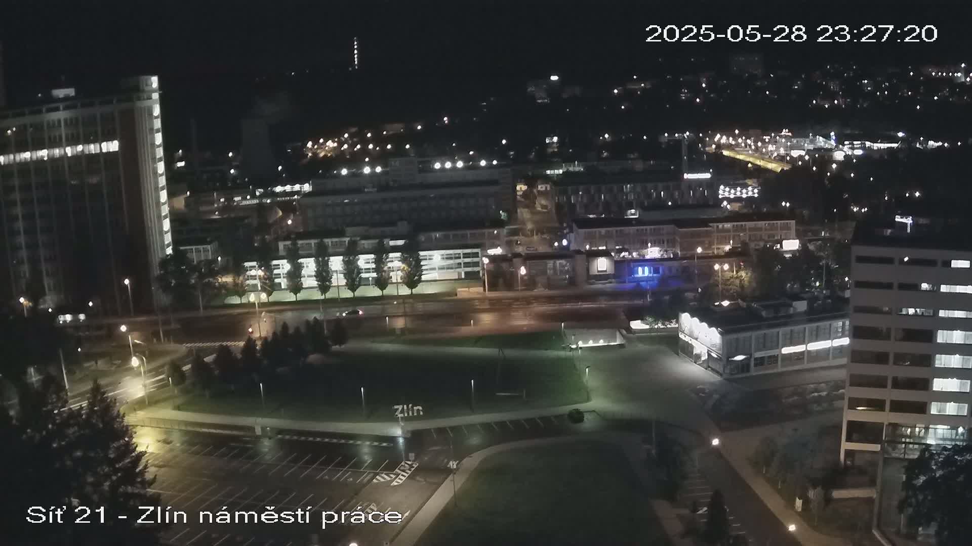 A nighttime aerial view of a city showing wet streets, illuminated buildings, and a grassy area, possibly a park.