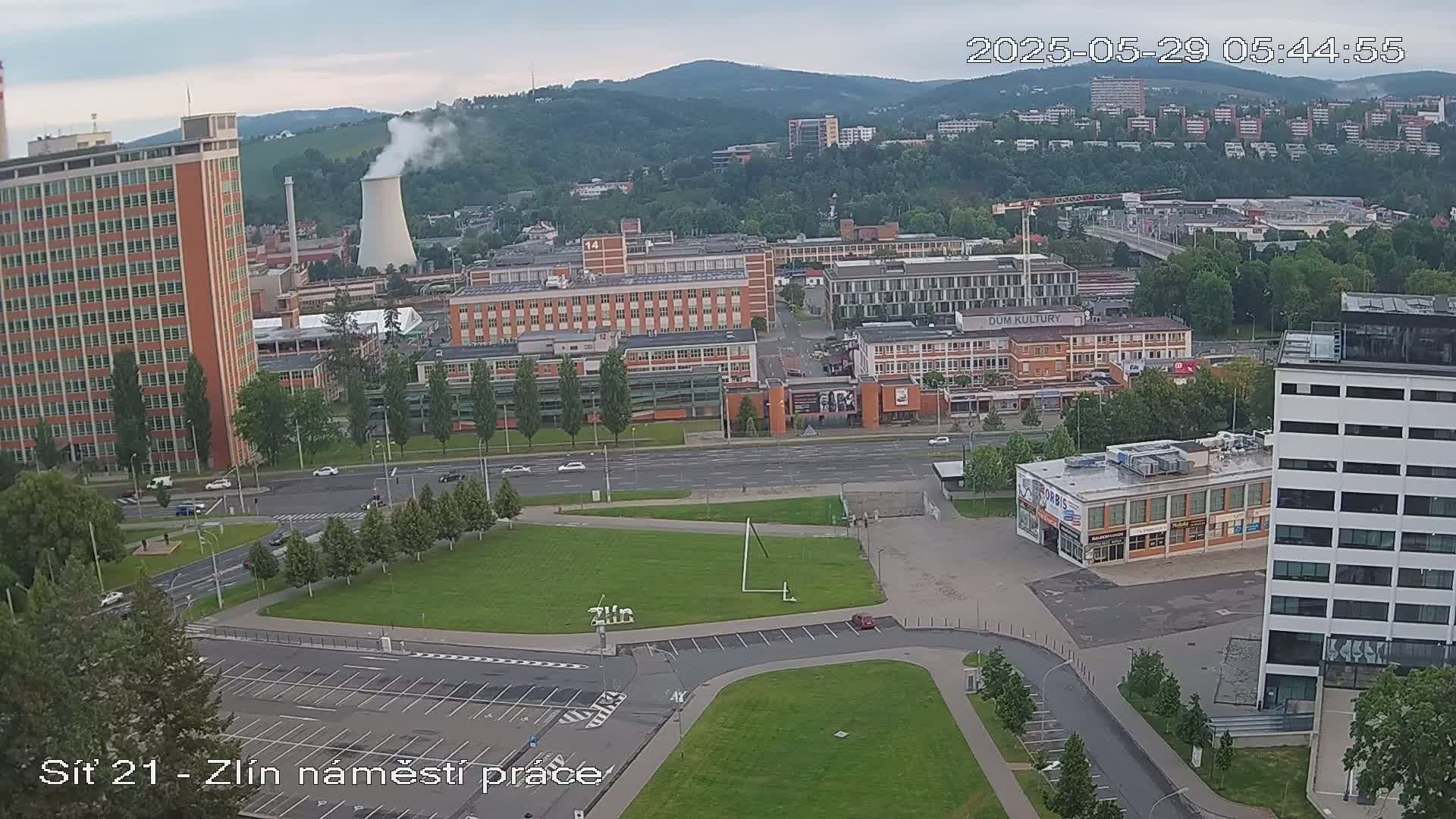 An overcast day shows a city scene with several buildings, including tall apartment blocks and industrial structures, a large grassy area, and a road with light traffic, all nestled against a backdrop of hills.
