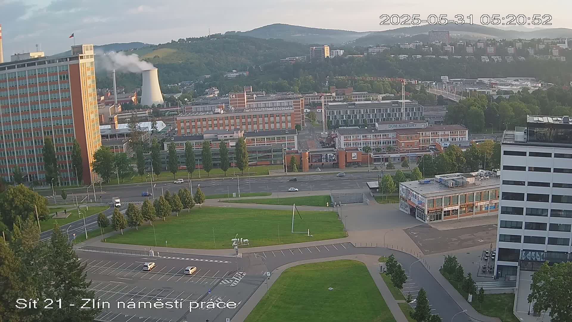 A high-angle, long shot of a city scene on a slightly hazy day, showing industrial buildings, a large grassy area, and several vehicles on roads and in parking lots.