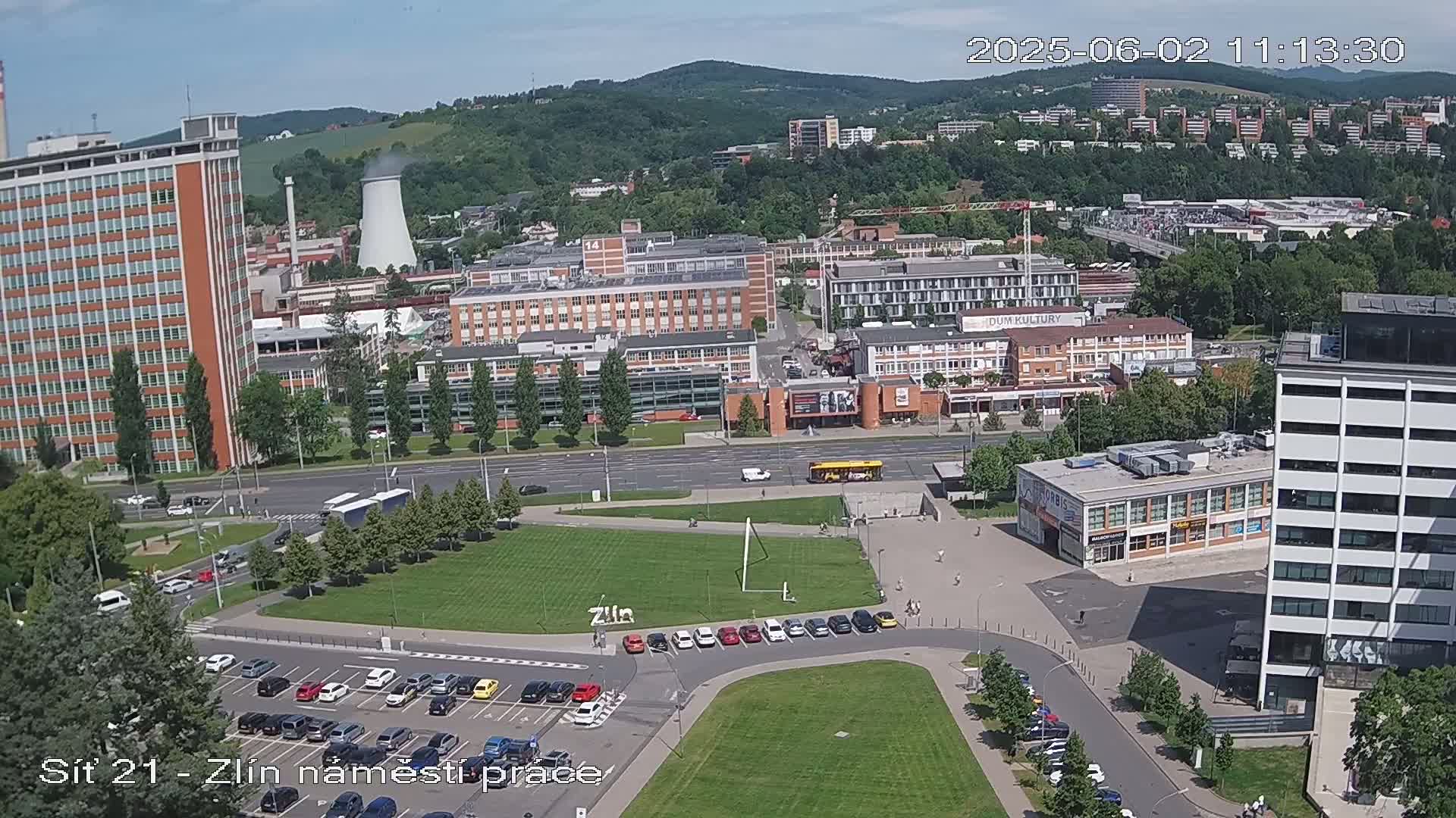 An aerial view of a city on a sunny day, showing a mix of industrial and residential buildings, streets with vehicles, and green spaces.