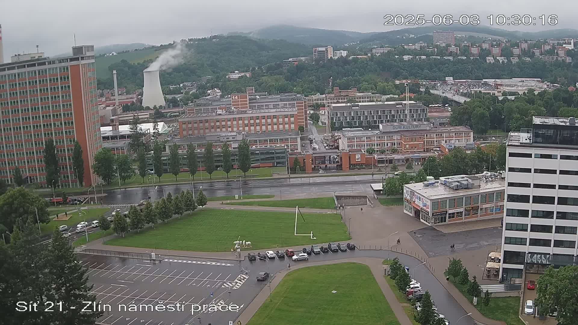 A high-angle, long shot shows a city landscape on an overcast day, featuring several buildings, a large grassy area, and cars parked in a lot.