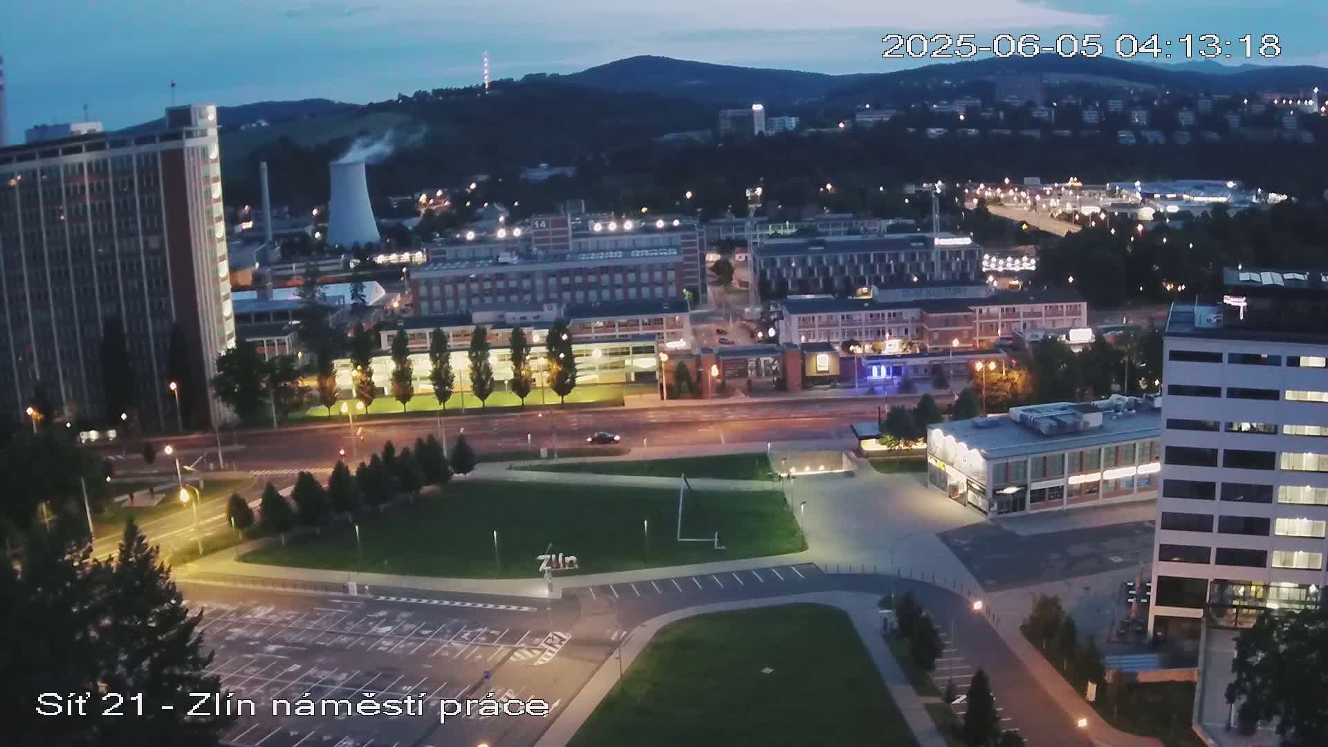 An aerial nighttime view of a city showing a large square with buildings and a parking lot, under a twilight sky.