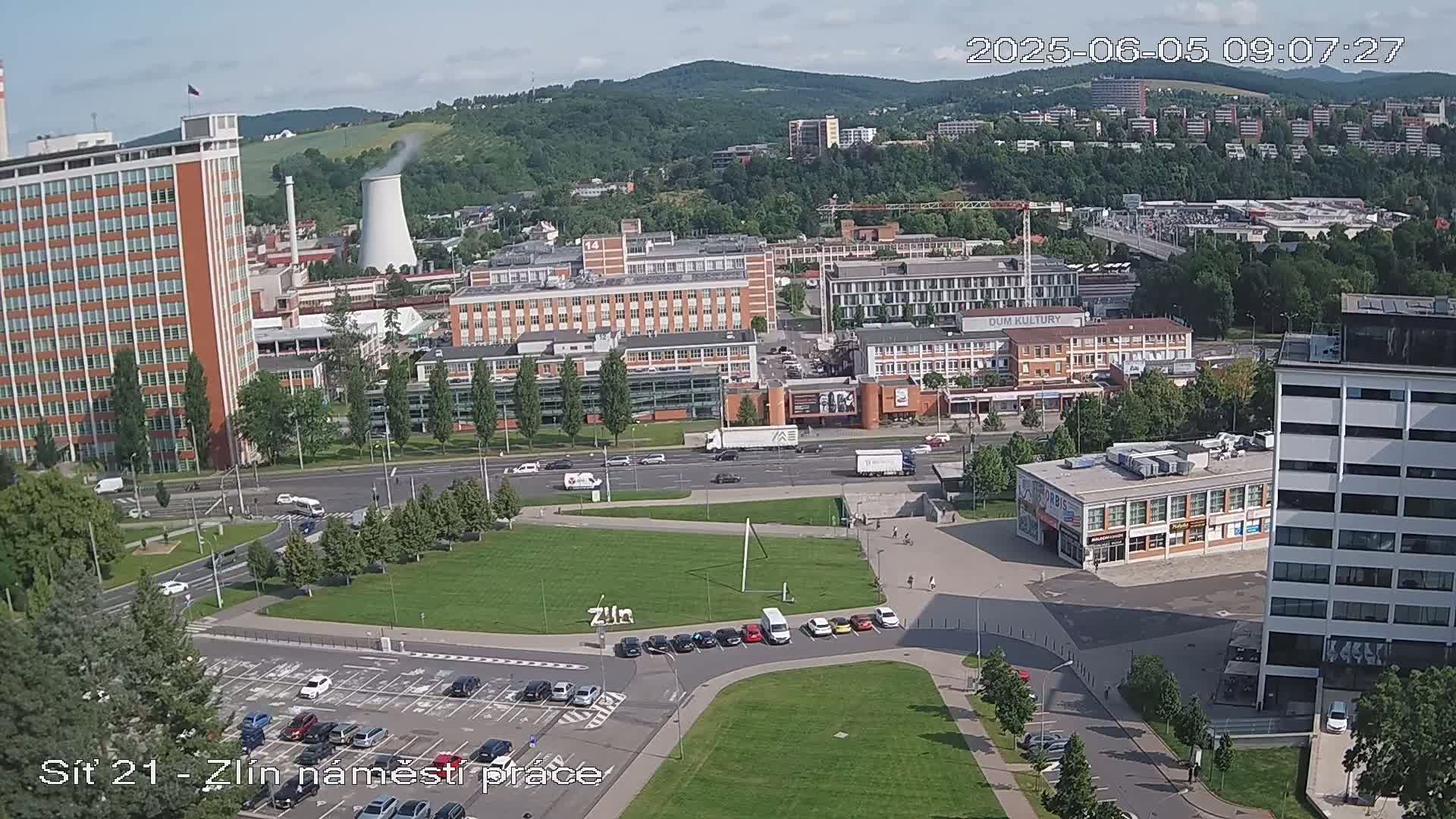 A high-angle, wide shot on a sunny day shows a city square with parked cars, buildings, and a grassy area, with a factory and cooling tower visible in the background.