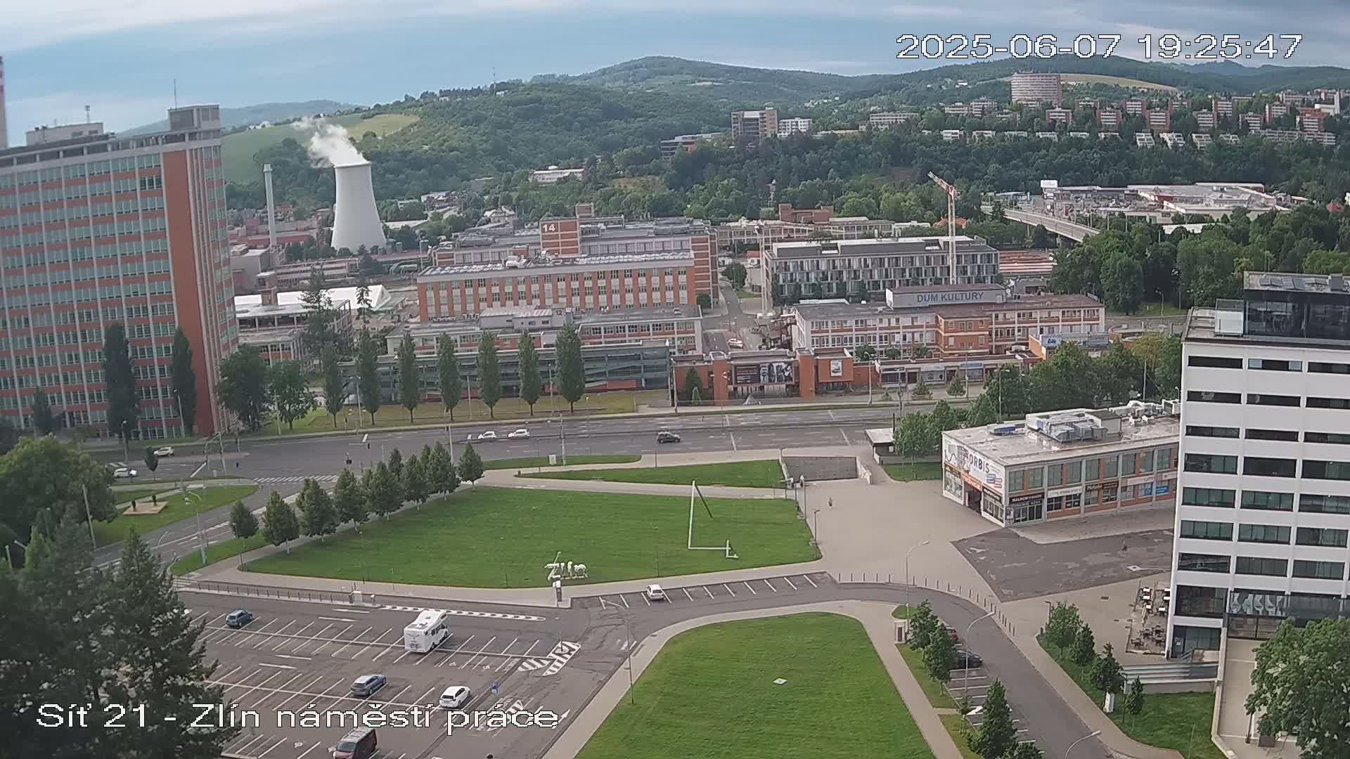 An aerial view of a city square with several buildings and a grassy area, under partly cloudy skies.