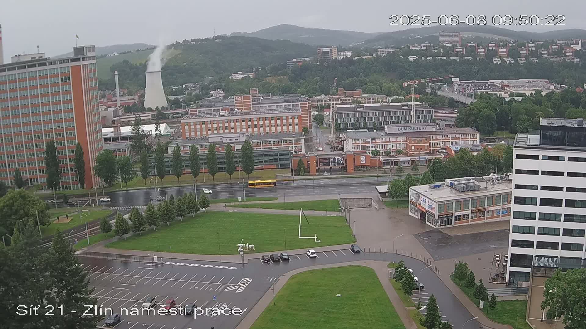 A high-angle, long shot on a rainy day shows a city with industrial buildings, apartment complexes, and a large grassy area with a parking lot in the foreground.