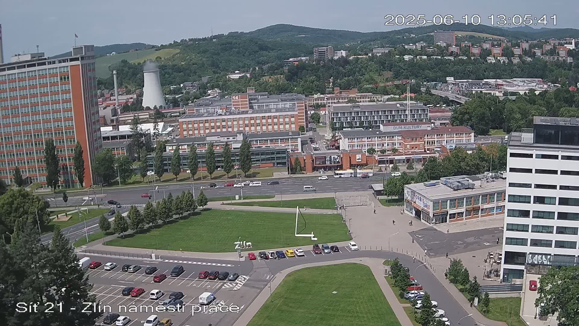 A high-angle, sunny view of a city's industrial and commercial area, showing buildings, streets, parking lots, and a grassy square.