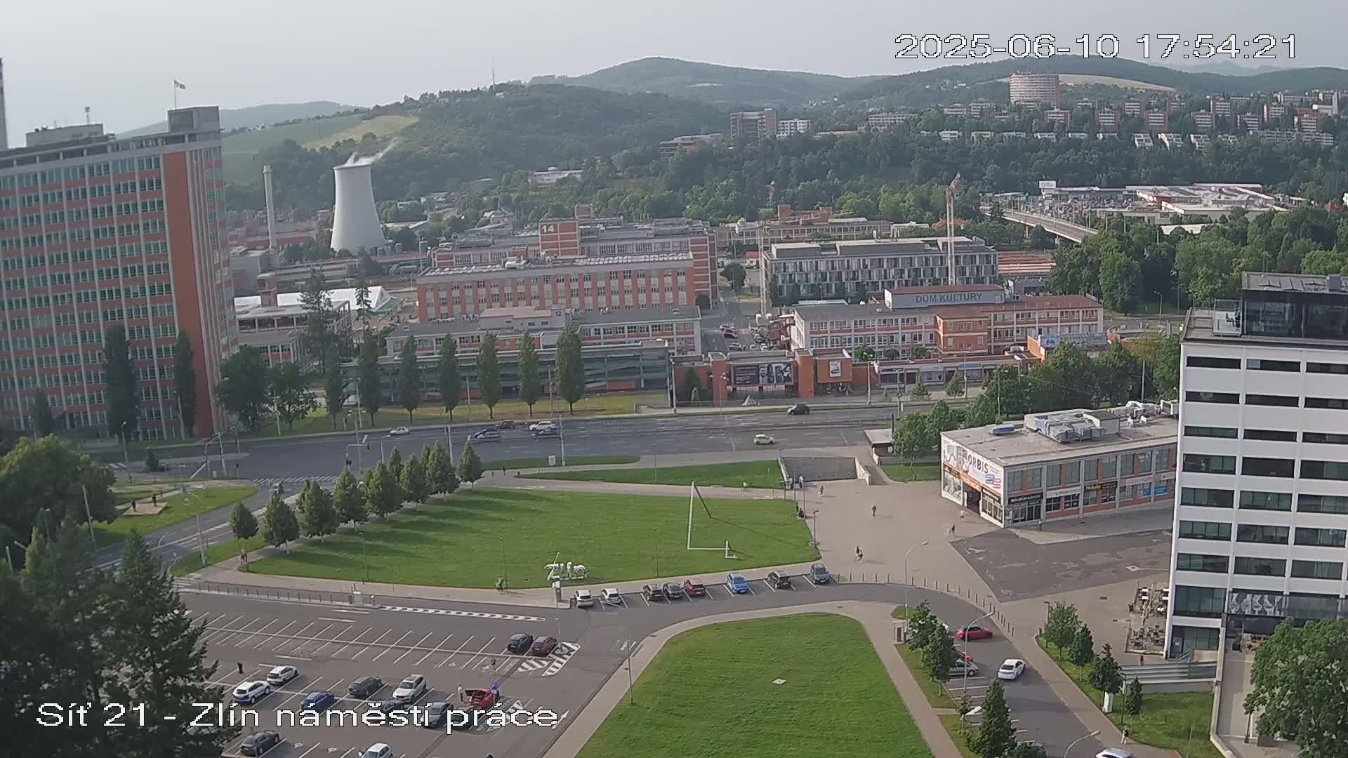 An aerial view of a city on a partly sunny day shows several buildings, streets, and a large grassy area with a parking lot.