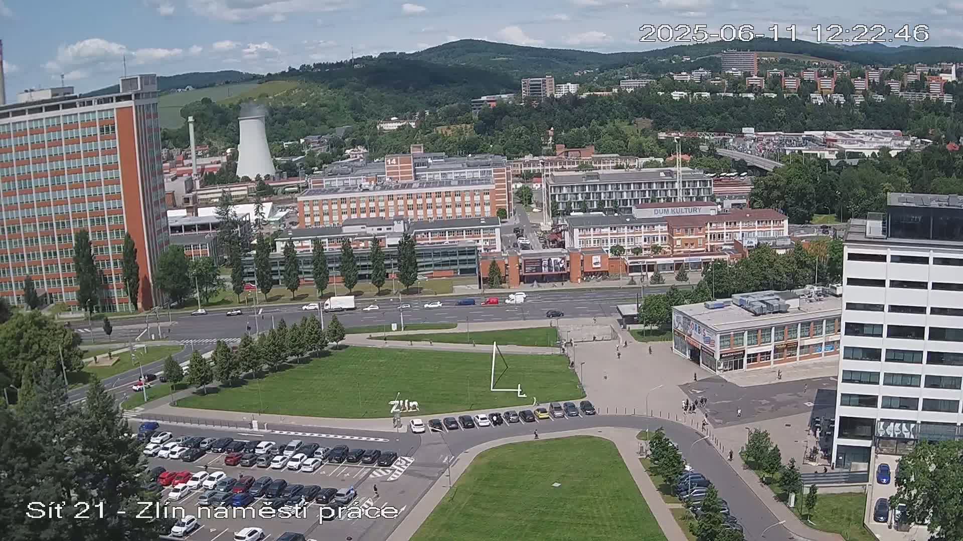 An aerial view on a sunny day shows a city square with parked cars, several multi-story buildings, and a grassy area, all surrounded by greenery and hills in the distance.