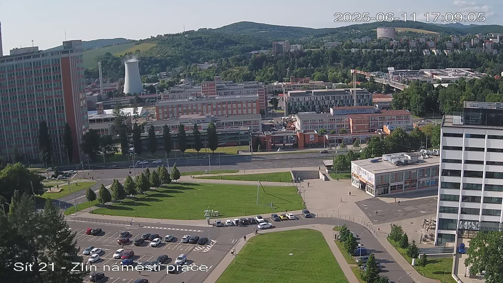 A high-angle, sunny view of a city with many buildings, roads, parking lots, and a large grassy area, partially obscured by trees in the background.