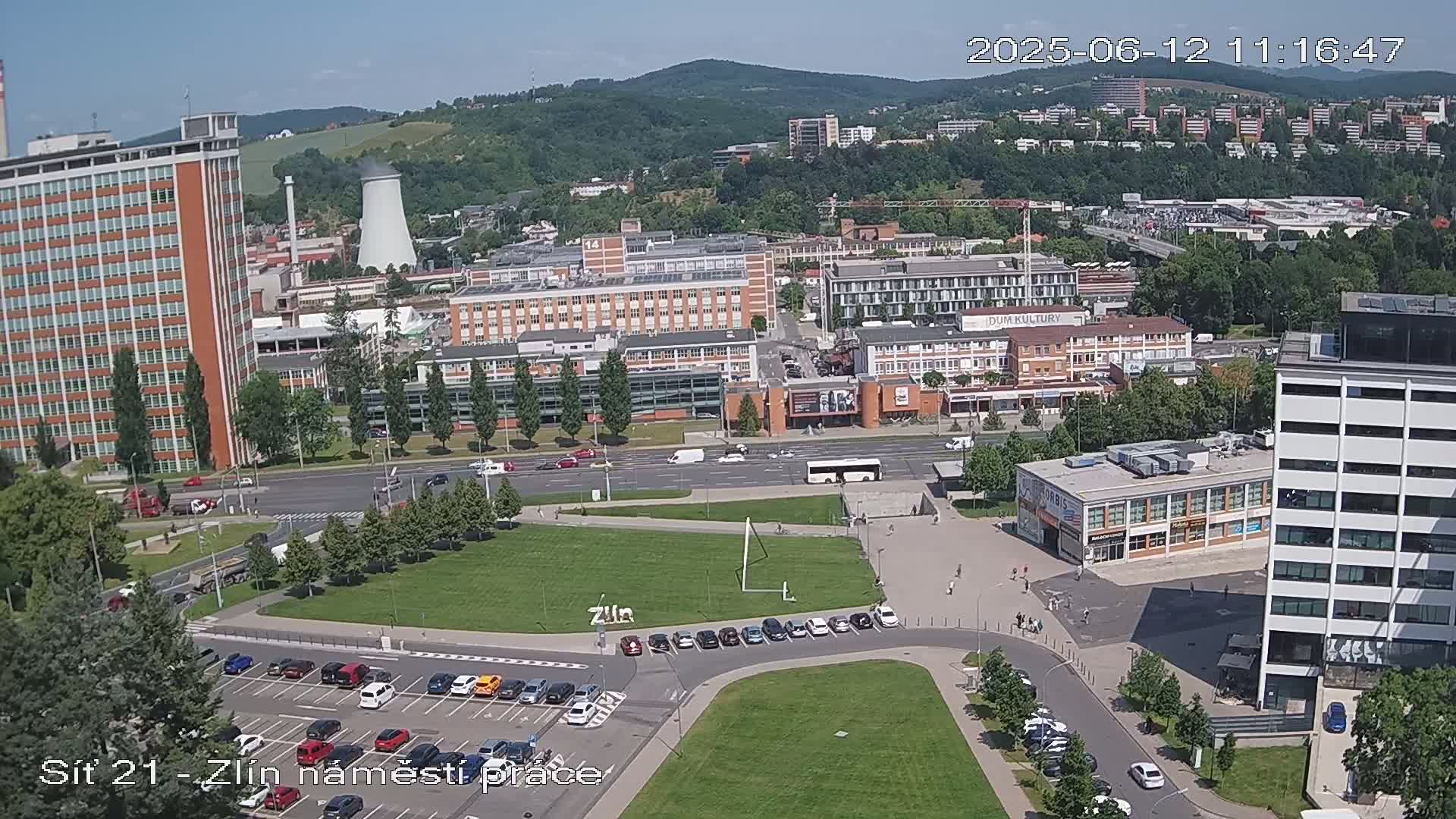 An aerial view of a city square on a sunny day, showing several buildings, parked cars, and a grassy area.