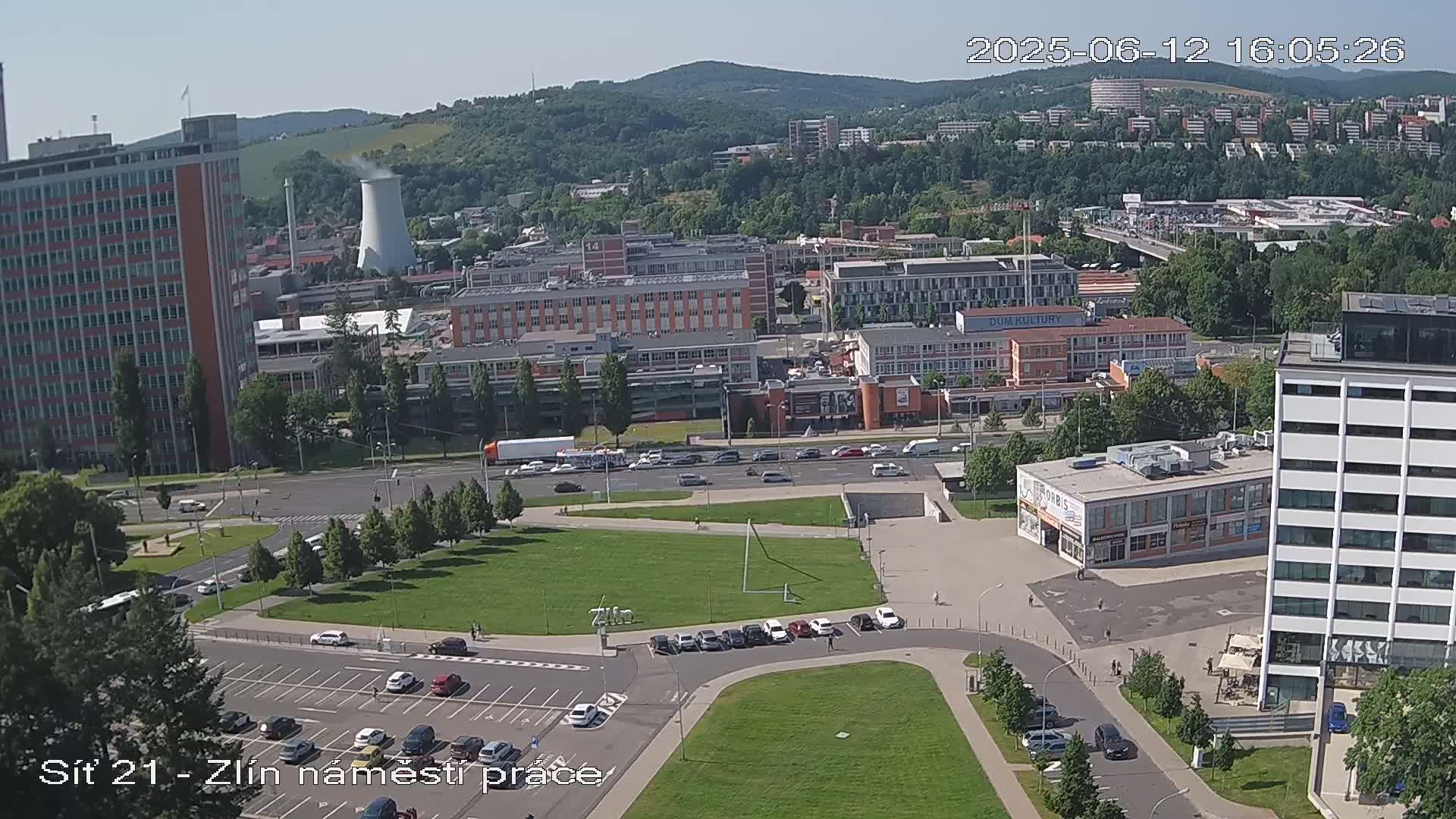 An aerial view of a city on a sunny day shows a large grassy area surrounded by buildings, parking lots, and roads, with some cars and people visible.