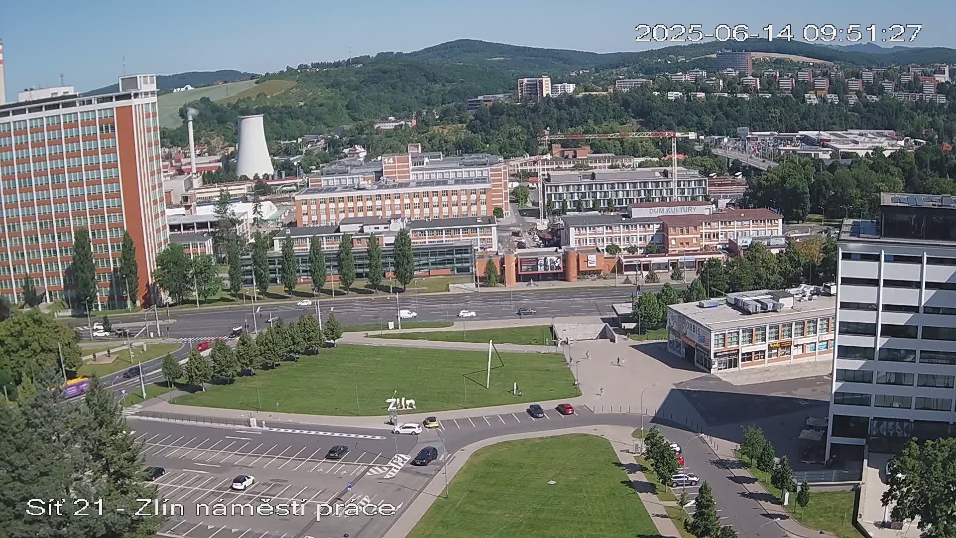 An aerial view of a city square on a sunny day, showing a variety of buildings, roads, and green spaces, with a few cars visible.