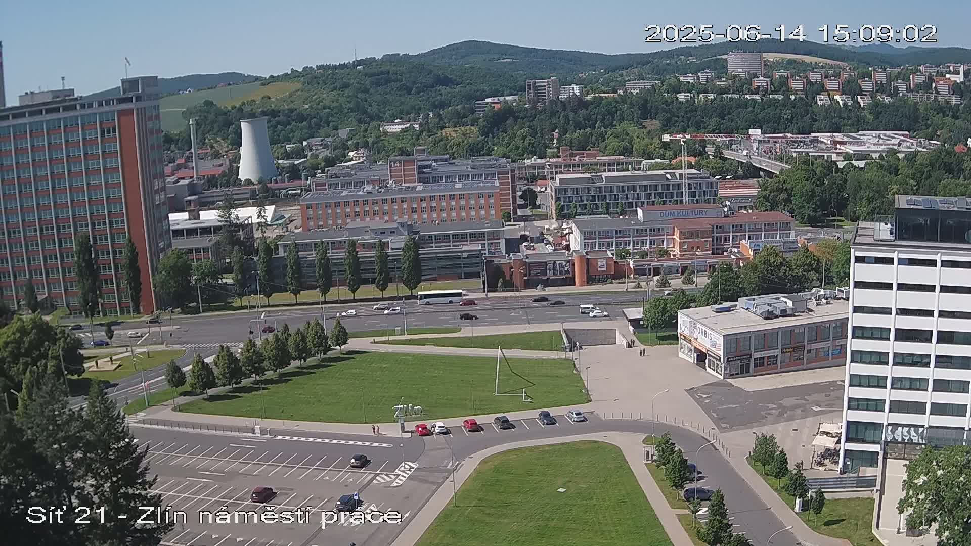 An aerial view of a city on a sunny day, showing several buildings, a large grassy area, and a roadway with cars.