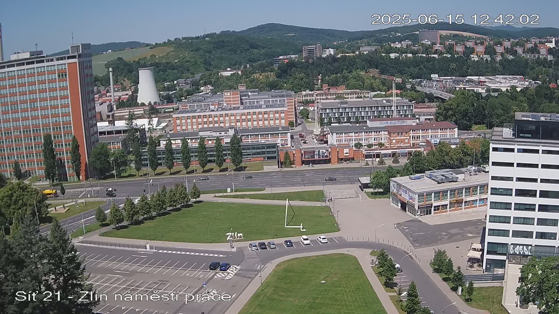 An aerial view on a sunny day shows a city with numerous buildings, including tall apartment blocks, industrial structures, and smaller commercial buildings, all surrounded by green spaces and roadways.