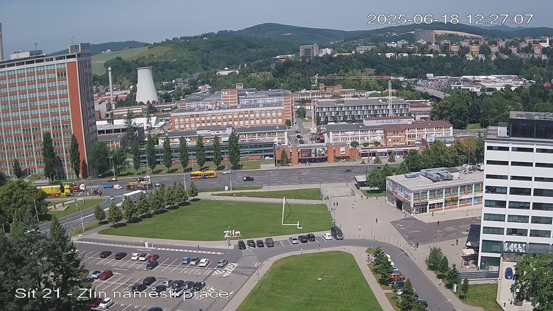 An aerial view of a city on a sunny day shows several buildings, streets with cars and buses, a grassy area, and a parking lot.