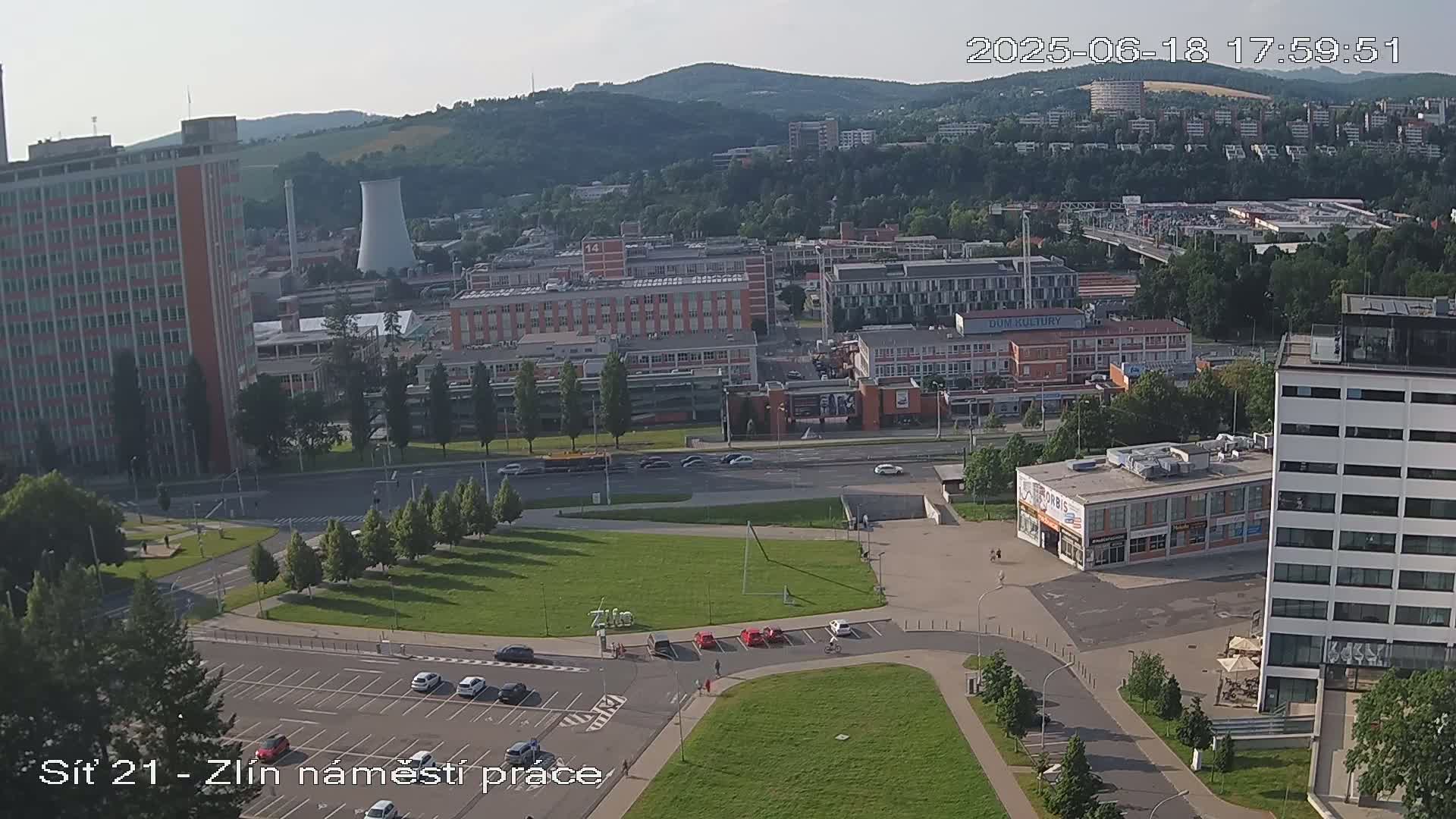 An aerial view of a city on a sunny day shows a large plaza with parked cars, surrounding buildings, and a few scattered trees.