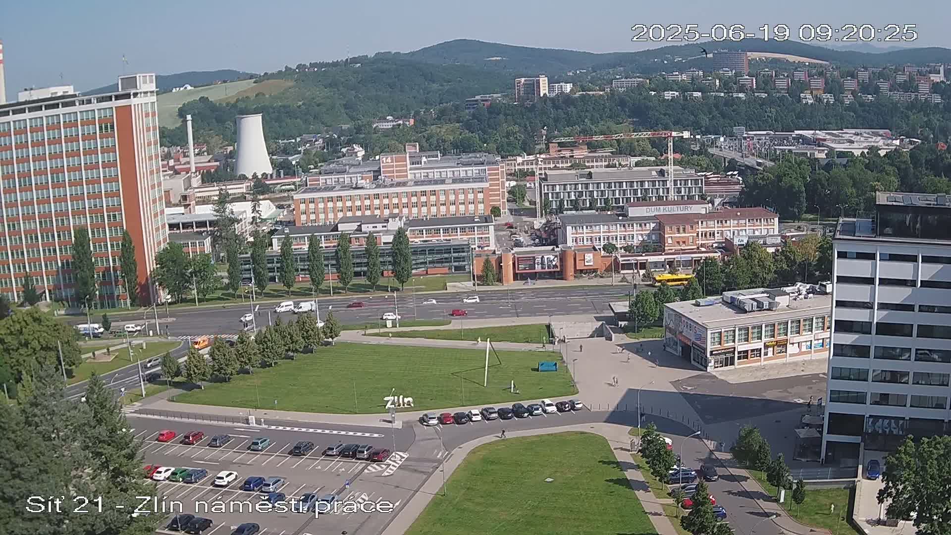 A high-angle, sunny view of a city with numerous buildings, a large grassy area, and several parked cars.