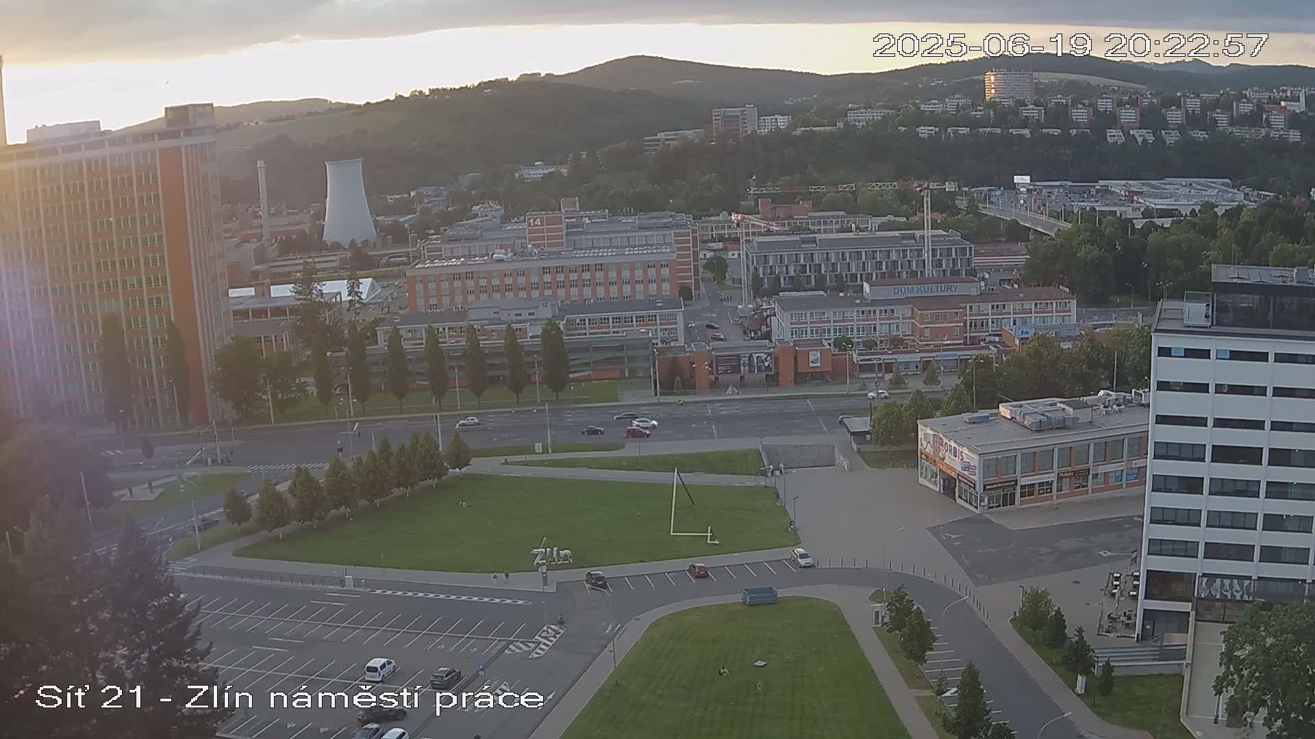An aerial view of a city at sunset, showing buildings, streets, and a grassy area with some cars parked.