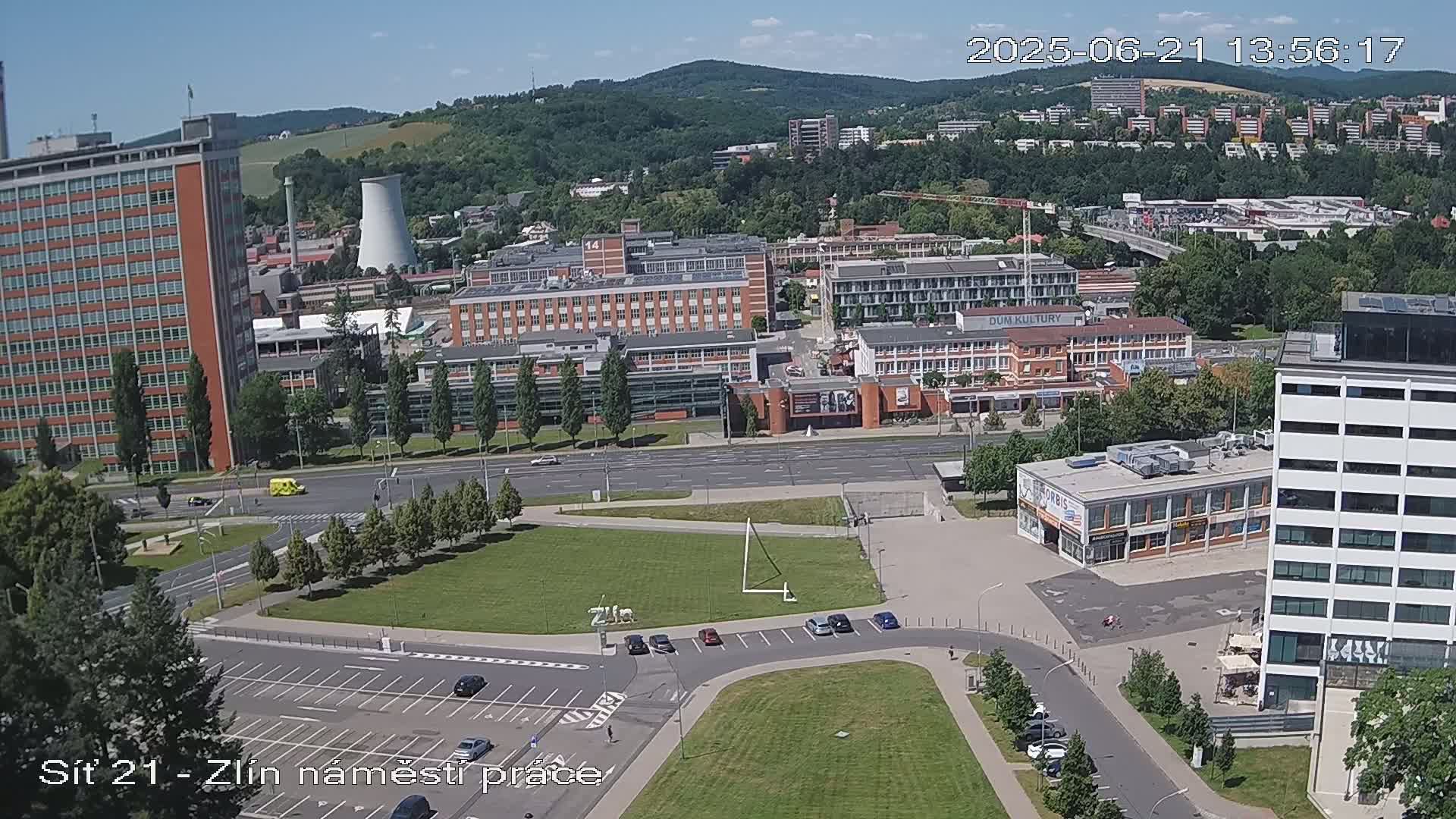 An aerial view shows a city scene on a sunny day, with numerous buildings, roads, and a large grassy area, including a parking lot.