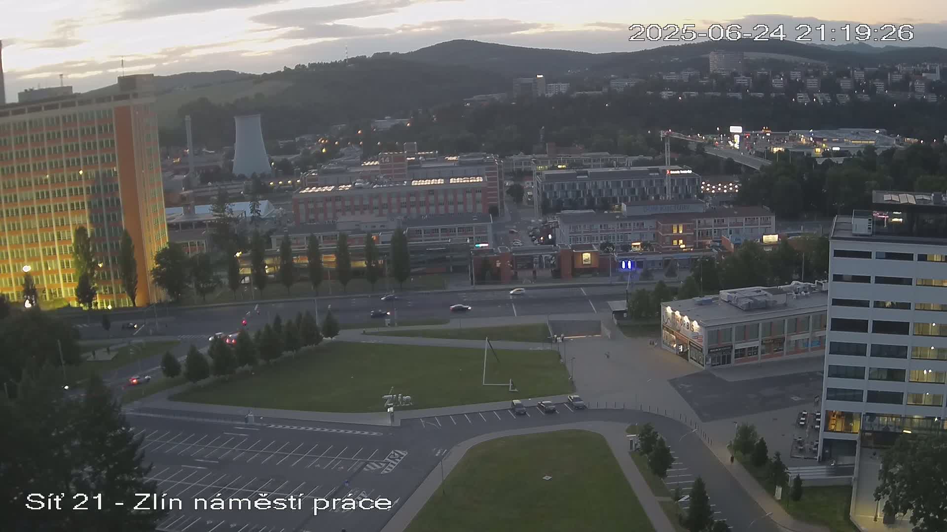 An elevated view of a city at dusk, showing buildings, roads with sparse traffic, and a grassy area, under a partly cloudy sky.