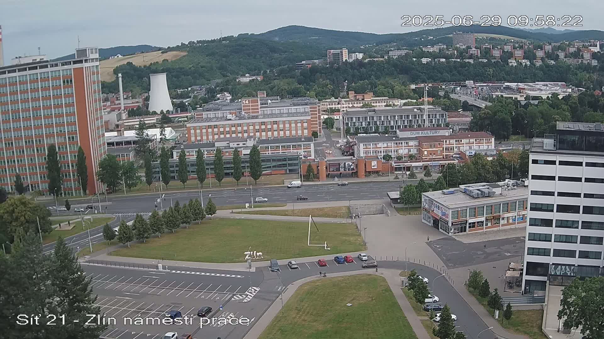 An aerial view of a city scene on an overcast day shows a large plaza with parked cars, surrounded by buildings of varying heights and styles, including a tall brick tower, and a wide road cutting through the area.