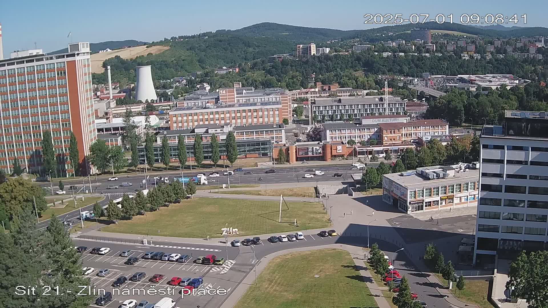 A sunny day reveals a city scene with numerous buildings, a large grassy area, and parked cars, all situated against a backdrop of rolling hills.