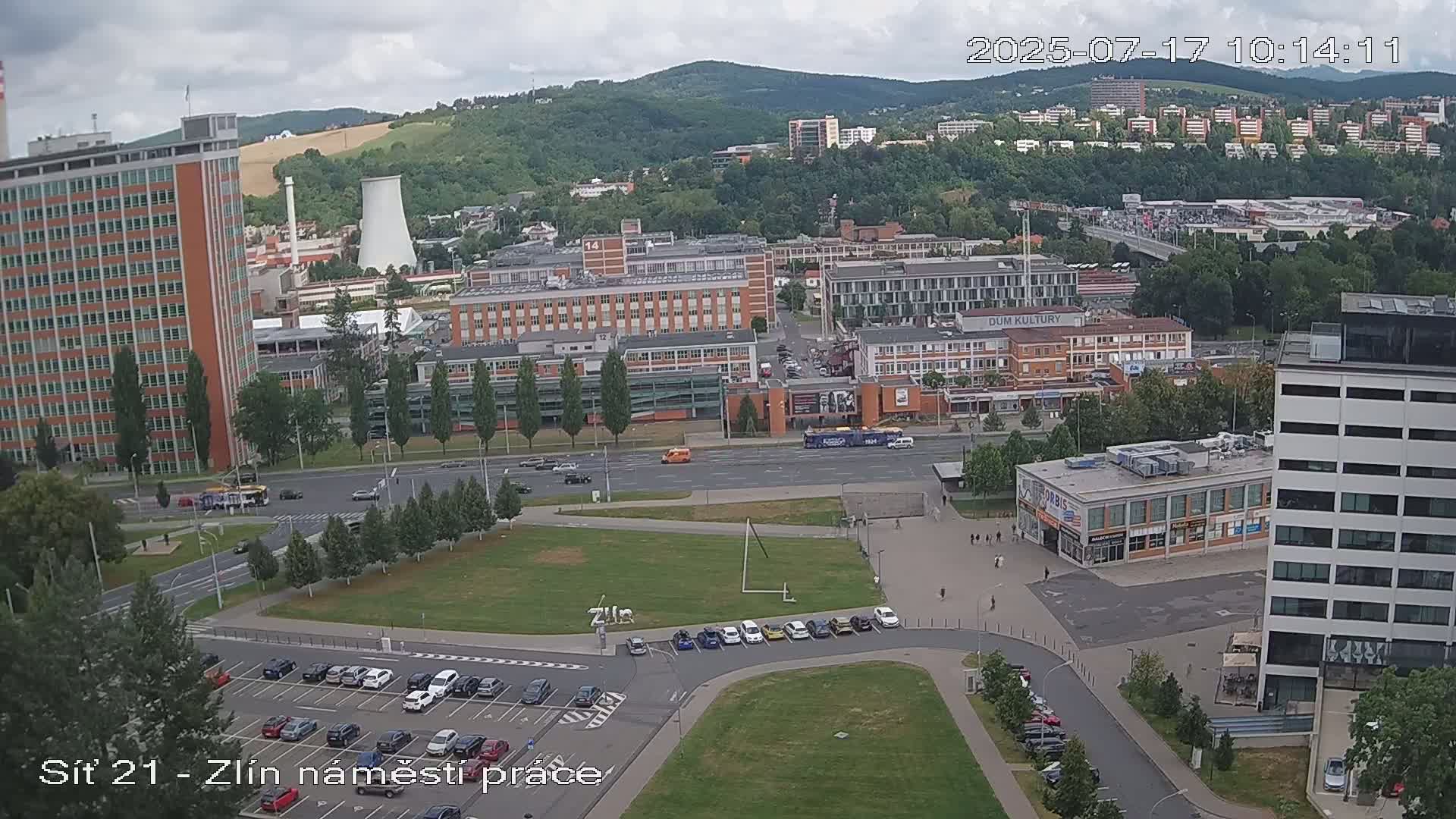 A high-angle, long shot of a city scene on a partly cloudy day, showing various buildings, streets, and a parking lot with cars, along with a grassy area.