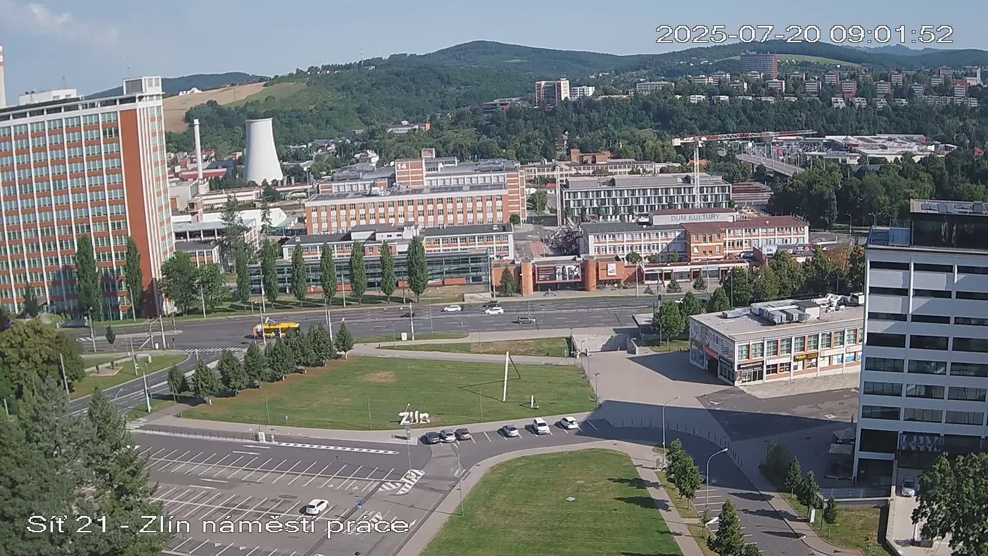 An aerial view of a city on a sunny day shows several buildings, a large grassy area, and a parking lot, with several cars parked.