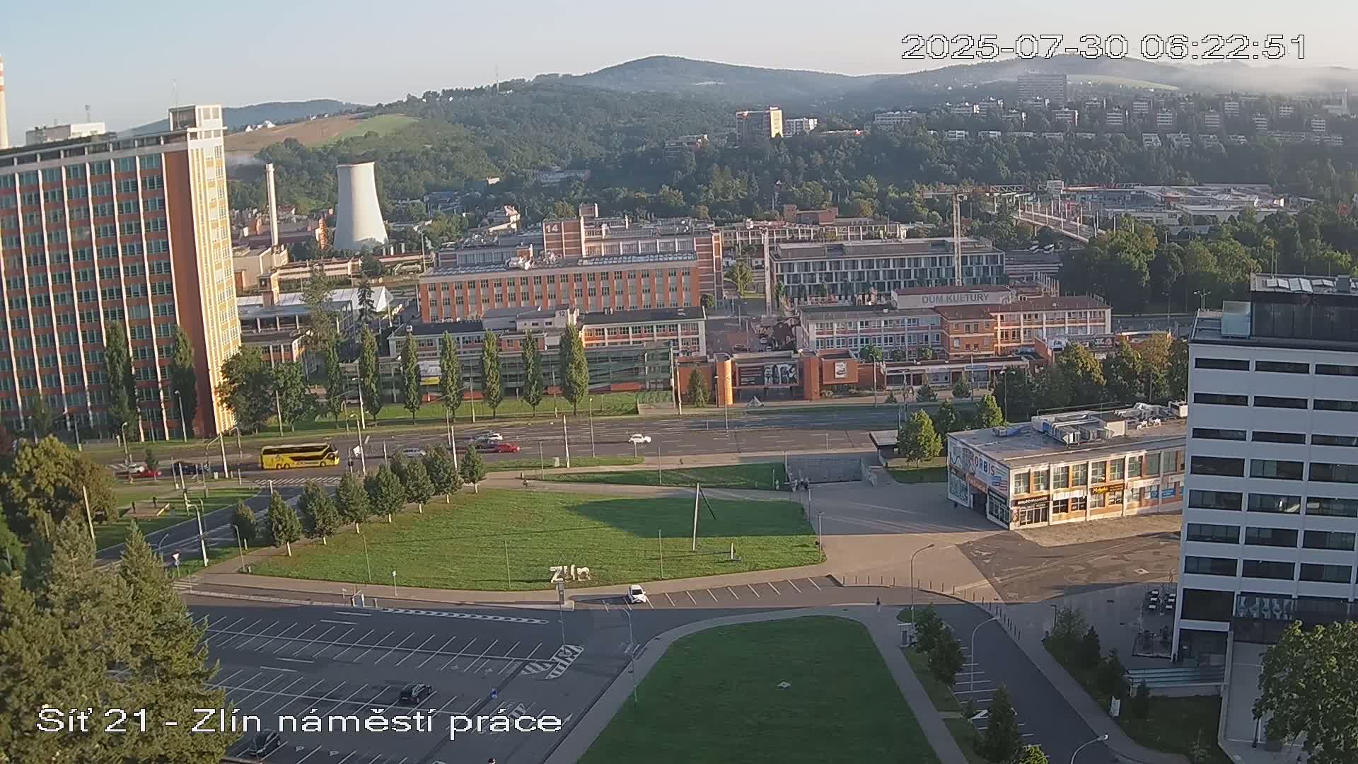An aerial view of a city on a sunny day shows several buildings, a large grassy area, and a parking lot, with several cars parked.