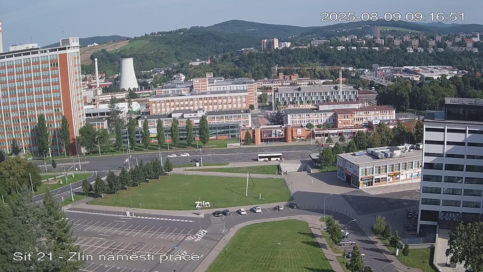 An aerial view on a sunny day shows a city scene with numerous buildings, a large grassy area, and a few vehicles, some of which are parked.