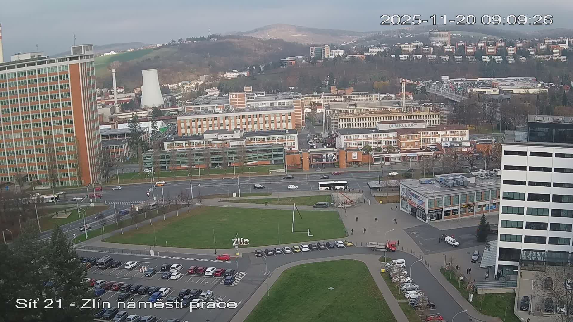 An elevated view of an urban landscape on an overcast day reveals a mix of tall and low-rise buildings, roads with vehicles, a central grassy square, and a large, full parking lot in the foreground, all framed by distant hills.