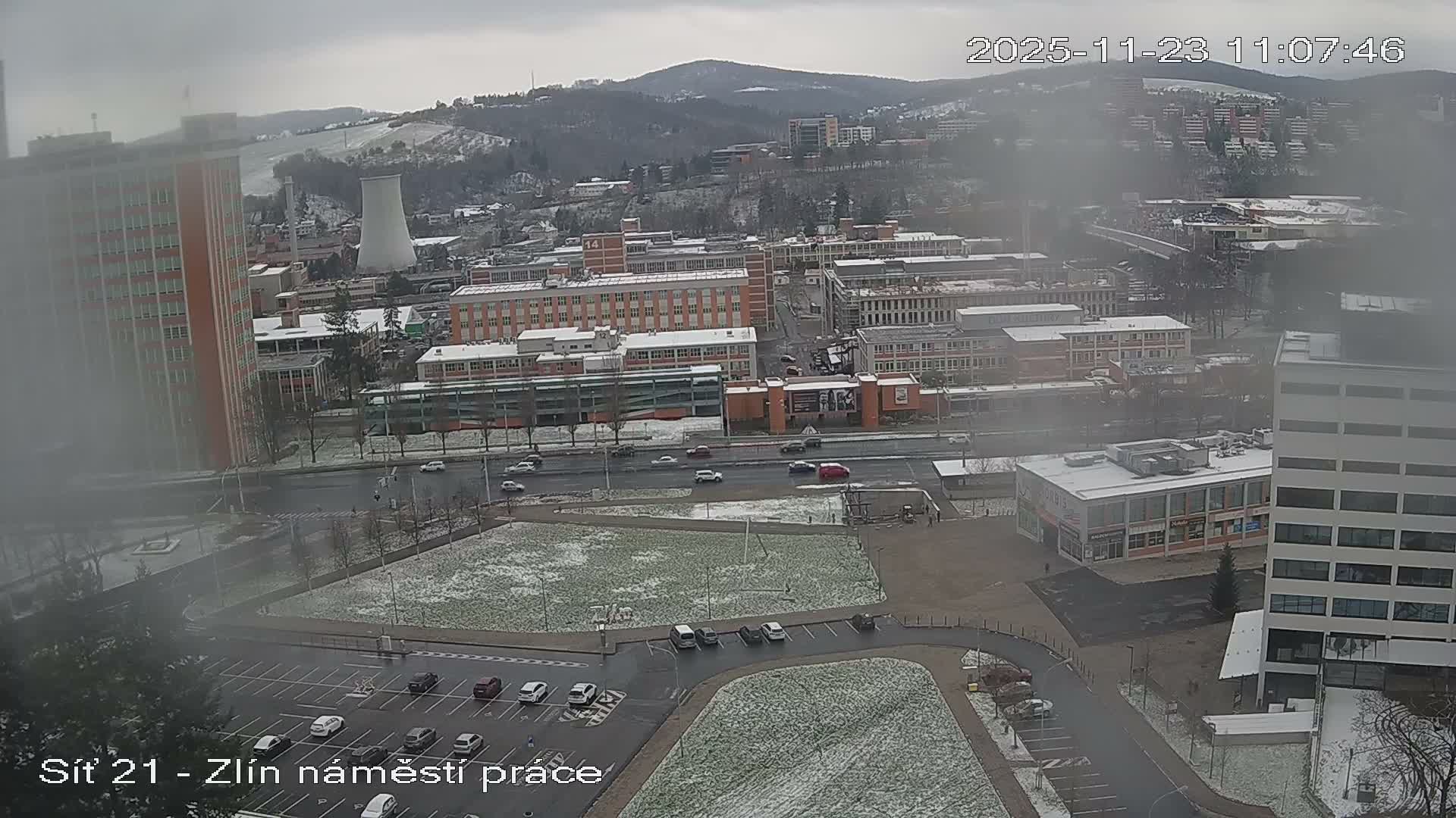 An overcast and hazy winter day overlooks a sprawling city scene featuring a prominent cooling tower, snow-dusted buildings and hills, moderate traffic on wet roads, and parked cars.