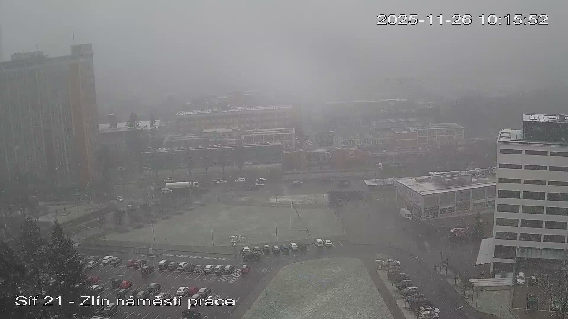 An elevated outdoor view reveals an urban landscape comprising numerous buildings, roads with vehicles, a large parking lot, and a sports field, all amidst heavy snowfall.