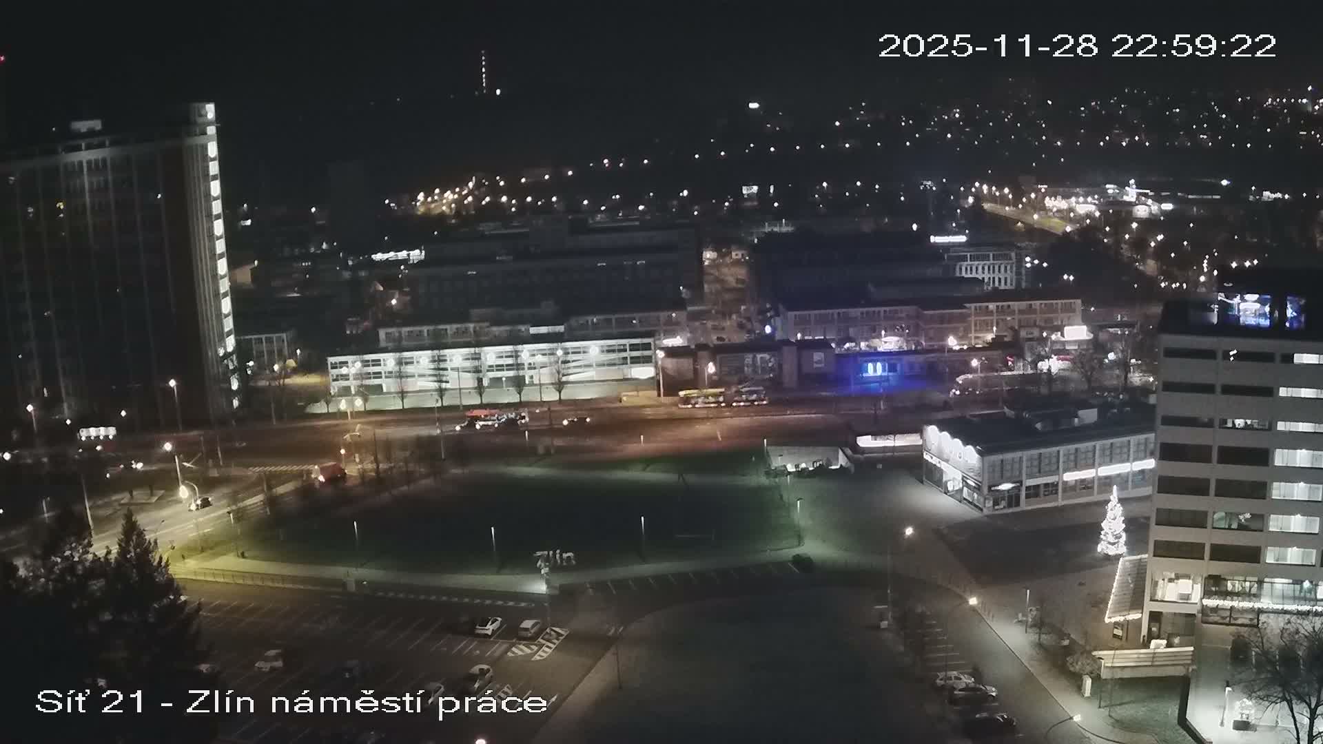 An aerial night view captures a clear urban landscape with illuminated buildings, busy streets with vehicles, and a brightly lit Christmas tree in a plaza.