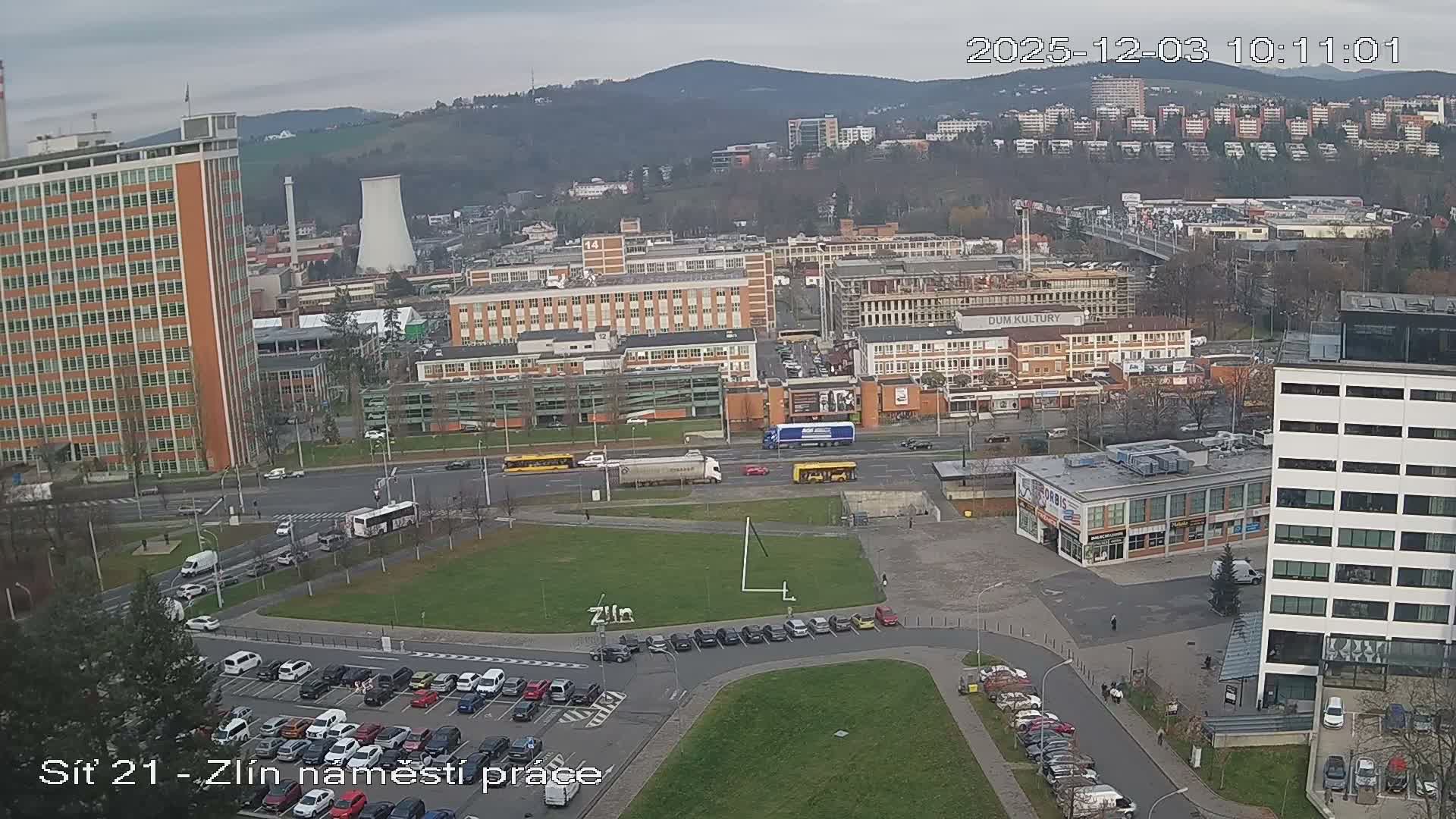 An overcast, grey day overlooks a bustling urban area with numerous buildings, busy roads filled with cars, trucks, and buses, multiple parking lots, and distant rolling hills.