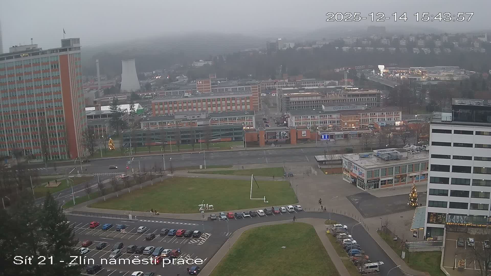 An overcast, grey day overlooks a bustling urban area with numerous buildings, busy roads filled with cars, trucks, and buses, multiple parking lots, and distant rolling hills.