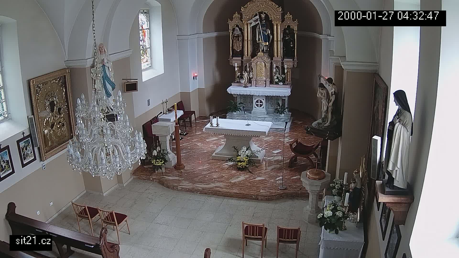 An indoor view of a church sanctuary shows a marble altar, several religious statues, a large crystal chandelier, and a few empty chairs.