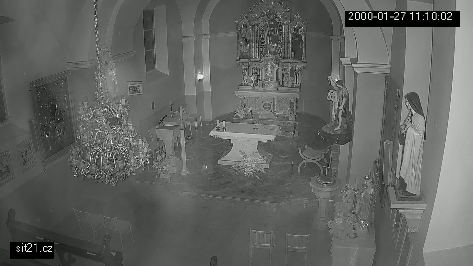 A nighttime, black and white, indoor view of a church sanctuary, showing an altar, several religious statues, a large chandelier, and empty chairs.