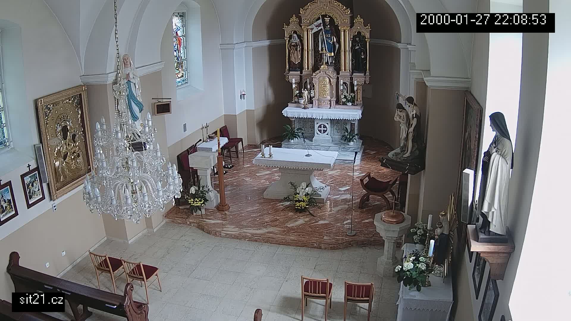 An indoor view of a church sanctuary shows an altar, several religious statues, a large crystal chandelier, and a few empty chairs.
