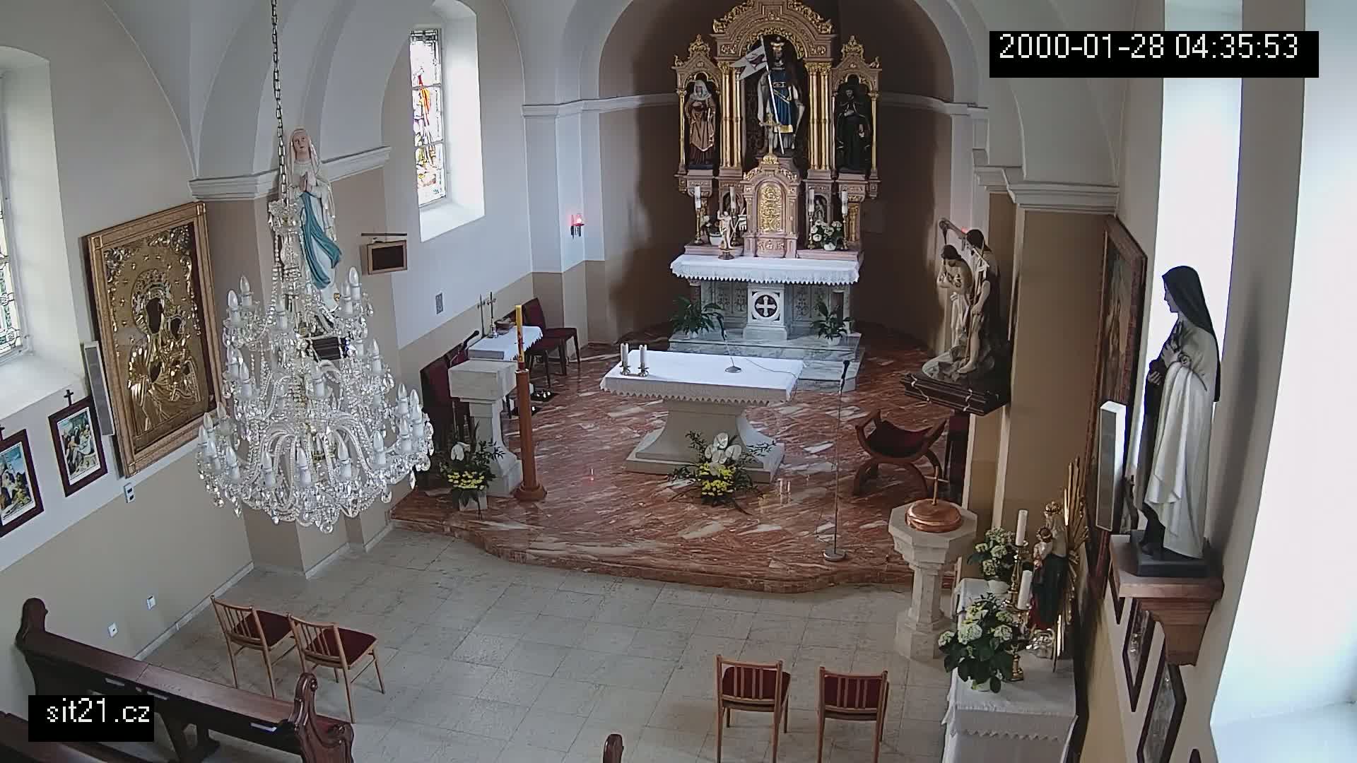 An interior view of a church, showing an altar, statues, a large crystal chandelier, and several chairs, with a marble floor.