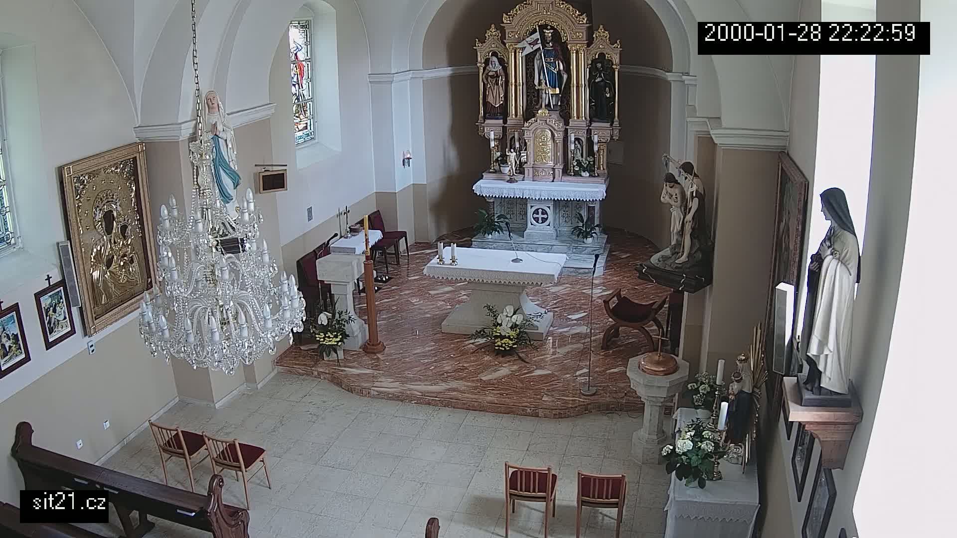 An indoor view of a church sanctuary shows an altar, several religious statues, a large chandelier, and empty chairs.