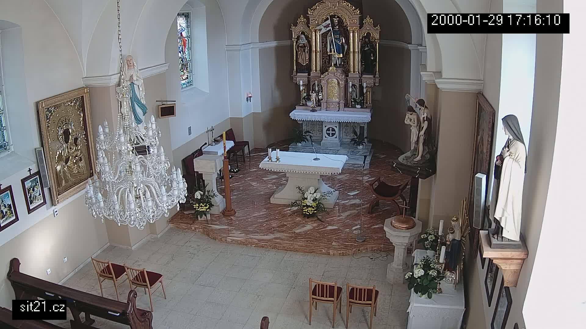 An indoor church sanctuary is shown with an altar, several religious statues and icons, a large crystal chandelier, and a few empty chairs.