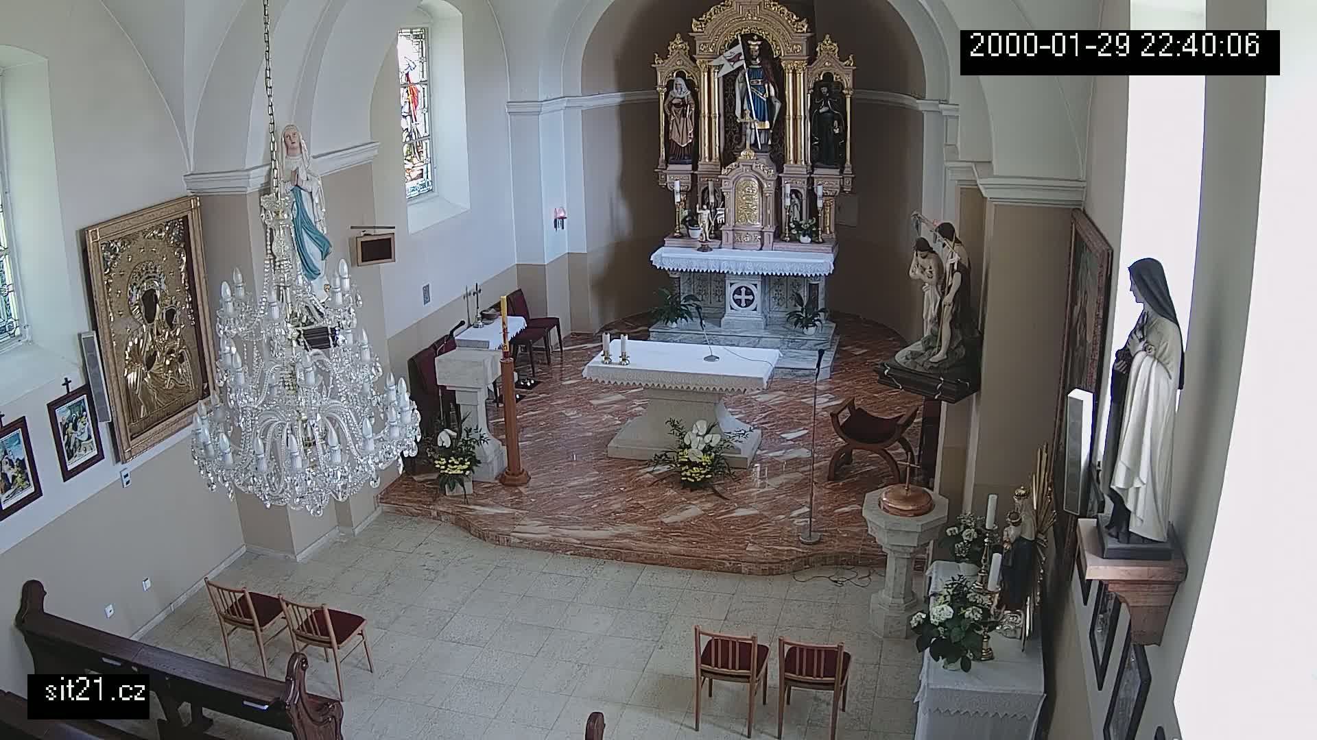 An interior view of a church shows an altar, several religious statues, a large crystal chandelier, and a few chairs.