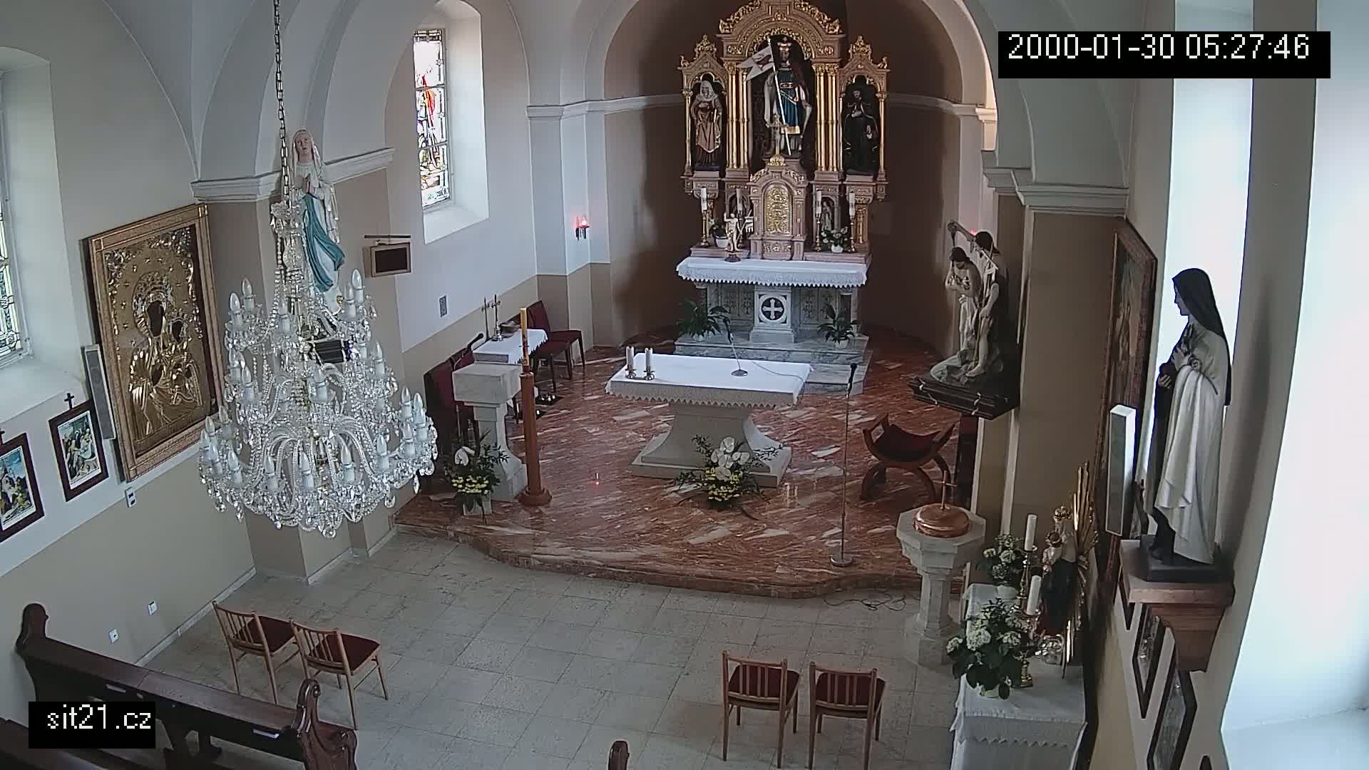 An indoor church sanctuary with marble floors, a large chandelier, several religious statues, and floral arrangements is shown.