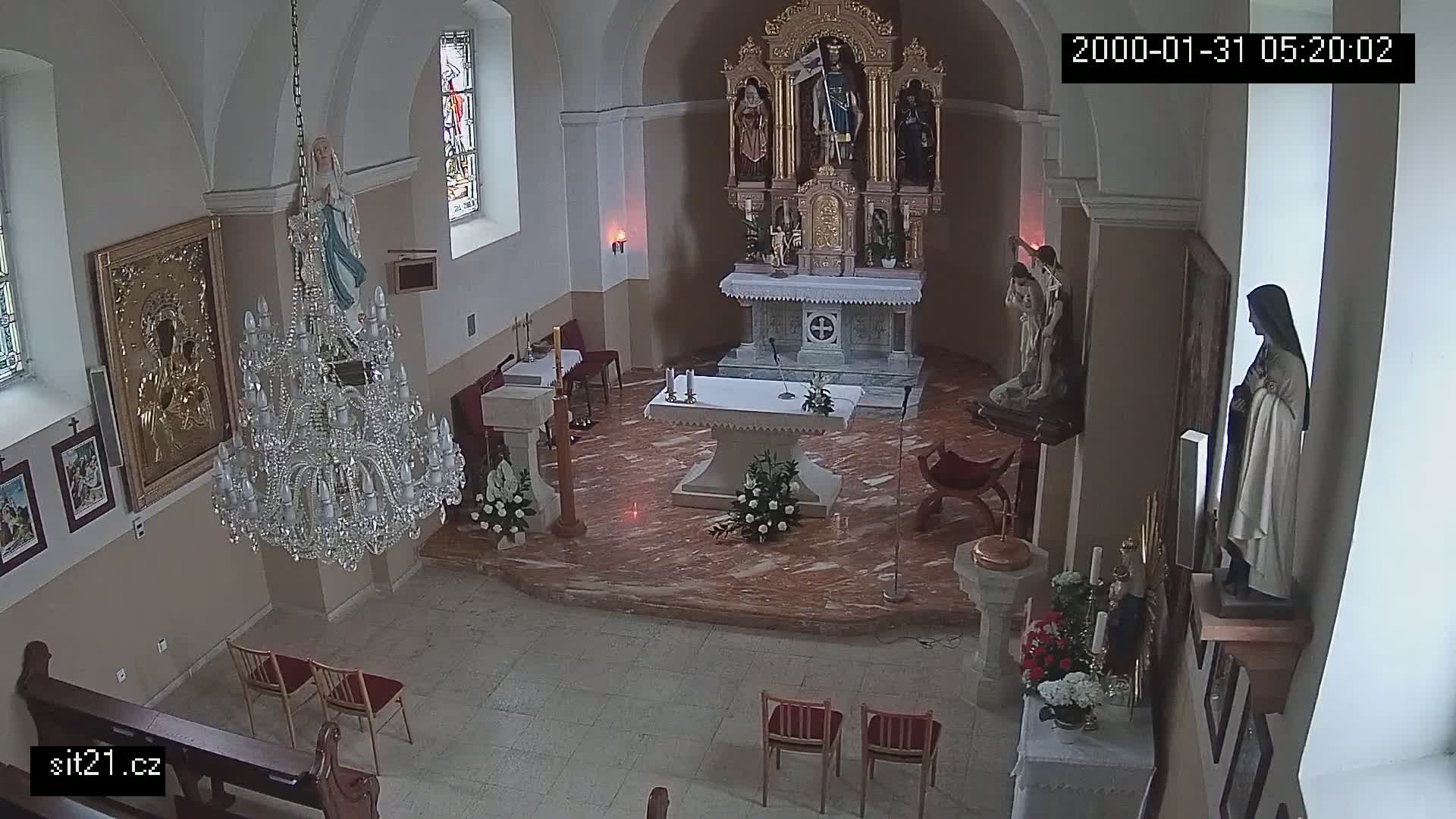 An indoor view of a church sanctuary, featuring a marble altar, religious statues, a crystal chandelier, and several chairs.