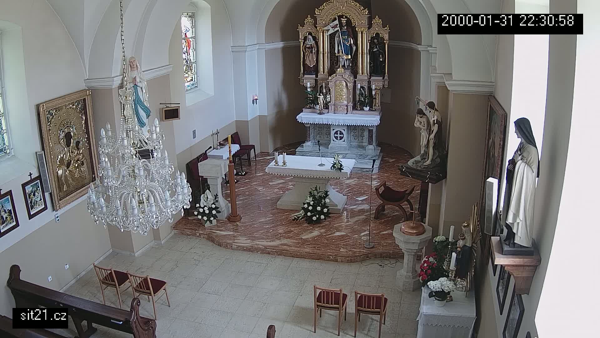 An indoor view of a church sanctuary with a marble floor, featuring an altar, statues, religious artwork, a chandelier, and a few chairs.