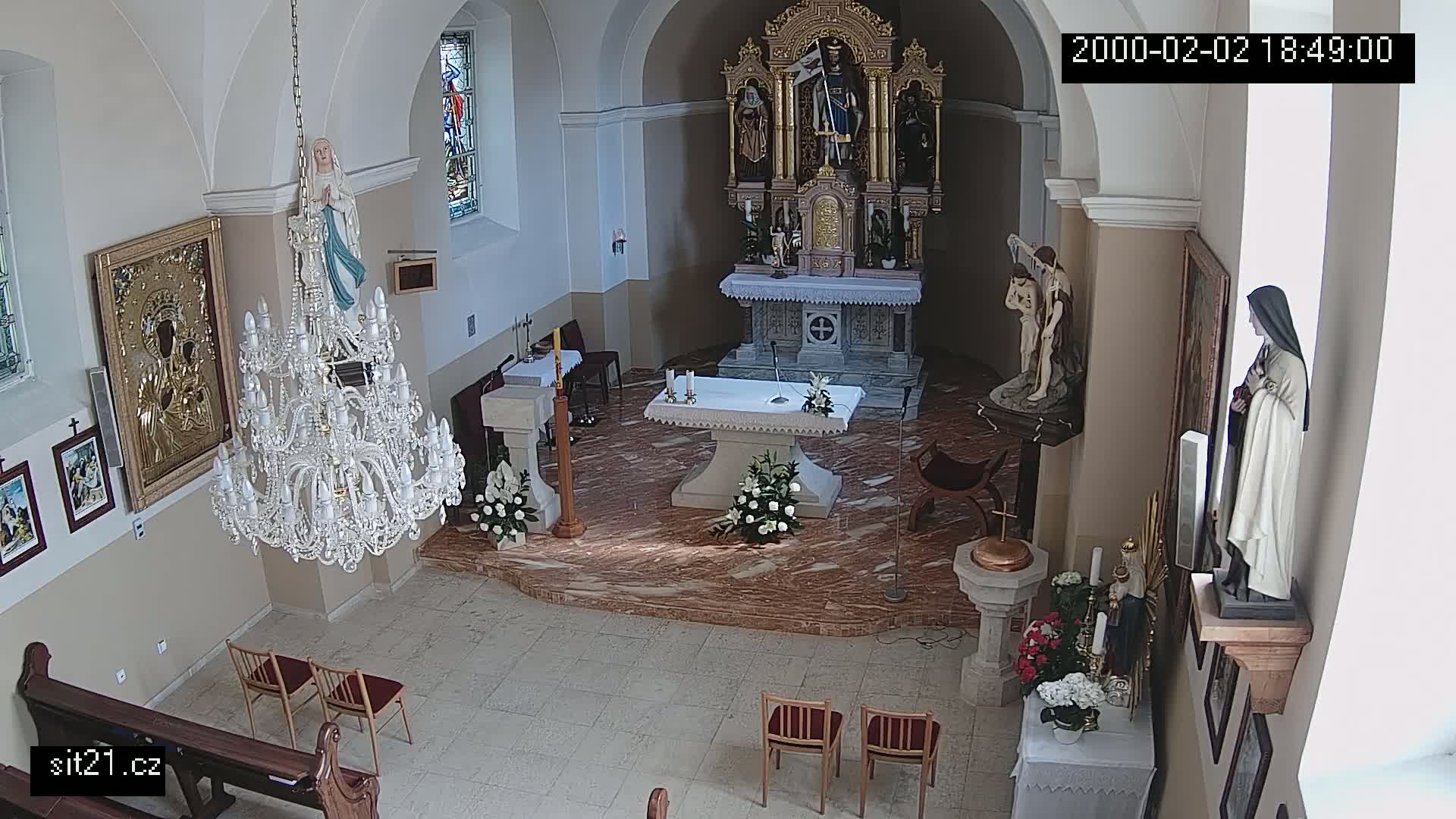 An indoor view of a church sanctuary shows an altar with floral arrangements, a large crystal chandelier, several religious statues and pictures, and a few chairs.