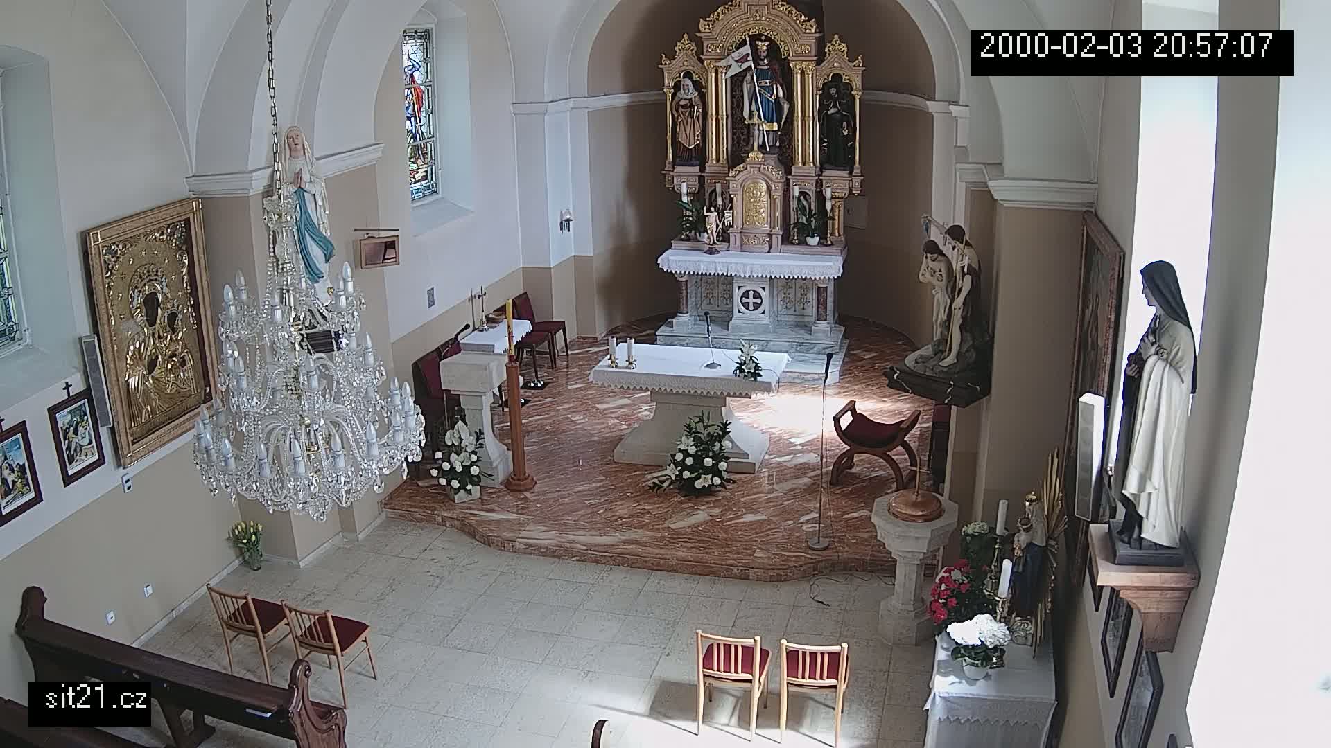An indoor view of a church sanctuary with an altar, several religious statues, a large chandelier, and floral arrangements, illuminated by natural light.