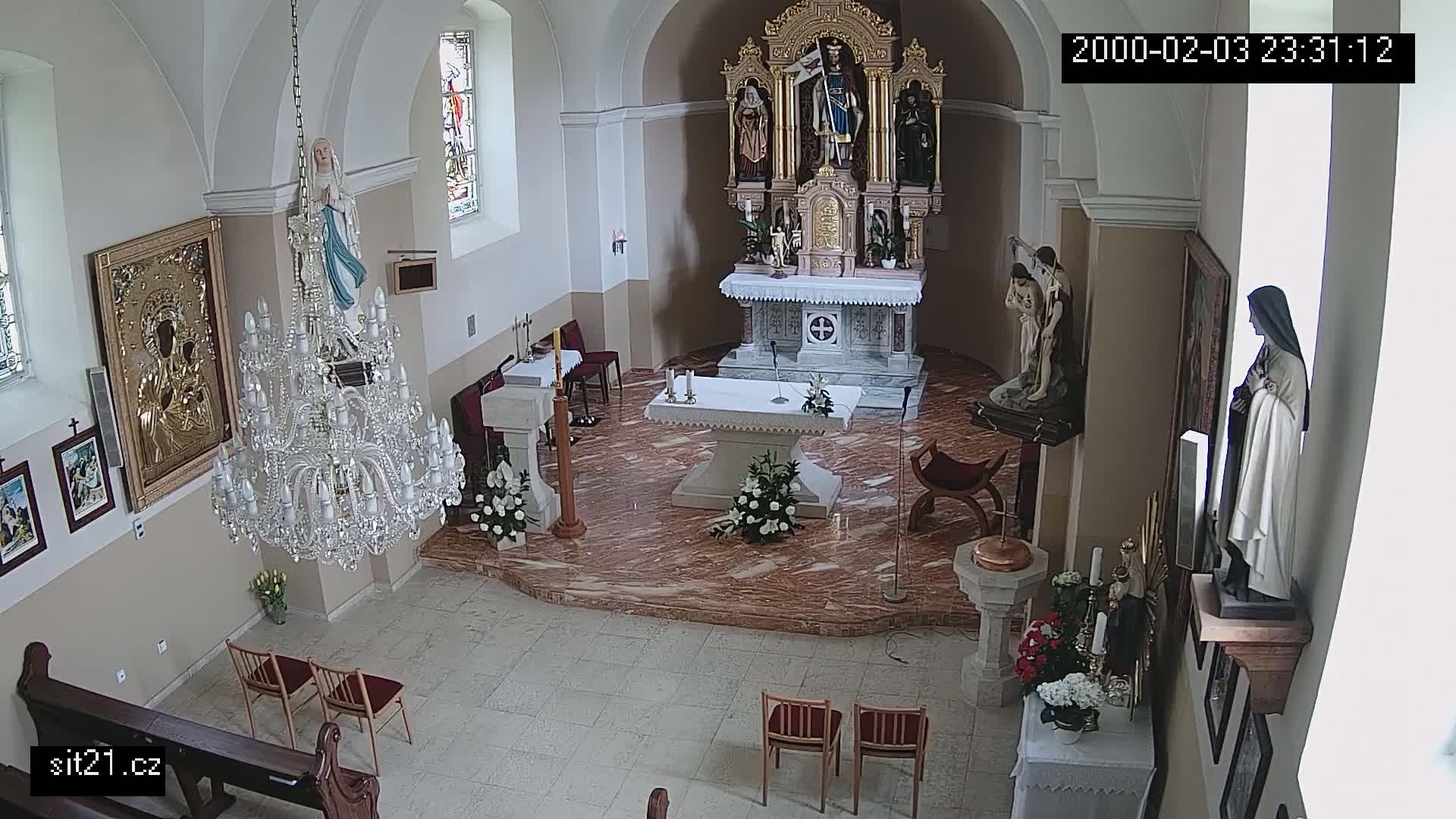 An indoor view of a church sanctuary with marble floors, an altar, several religious statues and pictures, a large crystal chandelier, and a few empty chairs.